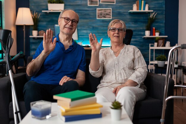 Portrait d'un couple de retraités assis sur un canapé en agitant les mains à la caméra dans le salon. Vieil homme et femme avec handicap de marche ayant des béquilles et un cadre de marche pour le transport