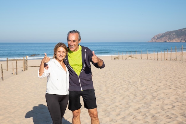 Photo gratuite portrait d'un couple de retraités profitant d'une belle journée active. homme et femme souriants après l'entraînement, debout, étreignant et montrant un bon geste pour la caméra. sport, mode de vie actif du concept de personnes âgées