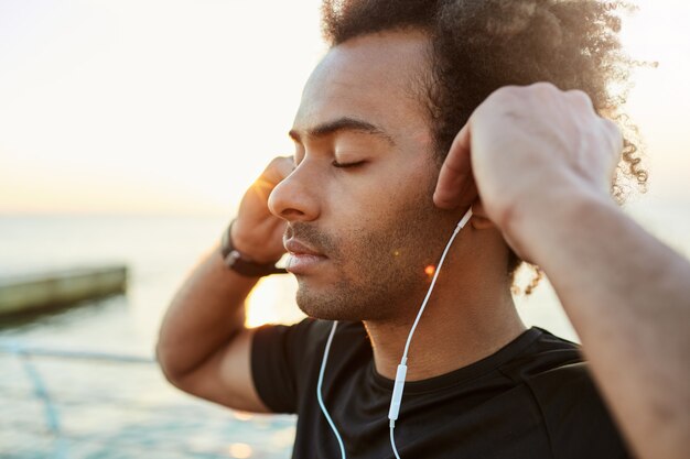 Portrait de coureur afro-américain médiateur et paisible avec une coiffure touffue et les yeux fermés écoutant de la musique. Tourné en plein air d'un sportif à la peau sombre en T-shirt noir se détendre après l'entraînement du matin ses