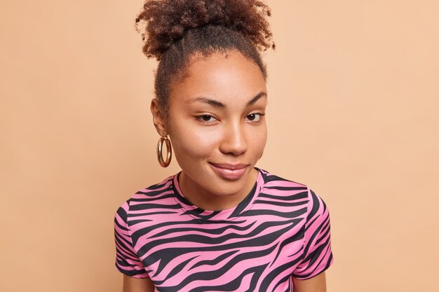 Portrait de curieuse belle femme regarde avec une expression sournoise à l'avant porte des boucles d'oreilles en t-shirt rayé regarde directement les poses avant contre le mur beige