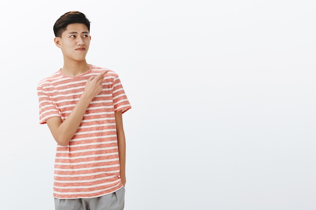 Portrait de curieux beau jeune mannequin asiatique en t-shirt rayé debout détendu sur un mur gris avec la main dans la poche à la recherche et pointant vers l'angle supérieur droit voir un espace copie intéressant