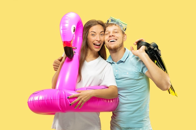 Portrait demi-longueur du beau jeune couple sur l'espace jaune. Femme et homme debout avec anneau de natation rose comme un flamant rose et des palmes