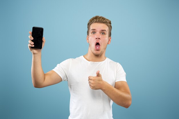 Portrait demi-longueur du jeune homme caucasien sur fond bleu studio. Beau modèle masculin en chemise. Concept d'émotions humaines, expression faciale, ventes, publicité. Affichage de l'écran du téléphone, paiement, paris.