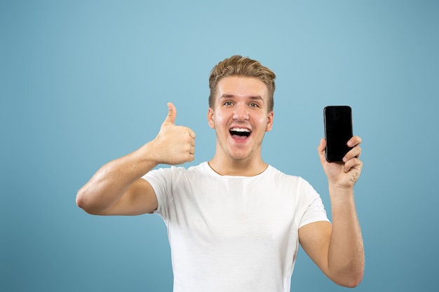 Portrait demi-longueur du jeune homme caucasien sur fond bleu studio. Beau modèle masculin en chemise. Concept d'émotions humaines, expression faciale, ventes, publicité. Affichage de l'écran du téléphone, paiement, paris.