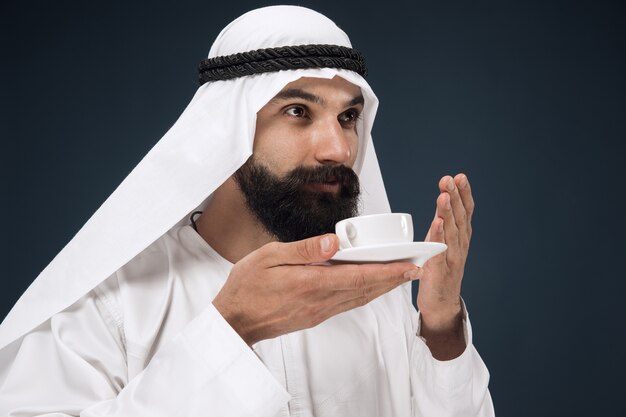 Portrait demi-longueur d'homme d'affaires saoudien arabe sur fond de studio bleu foncé. Jeune mannequin debout et buvant du café ou du thé. Concept d'entreprise, finance, expression faciale, émotions humaines.