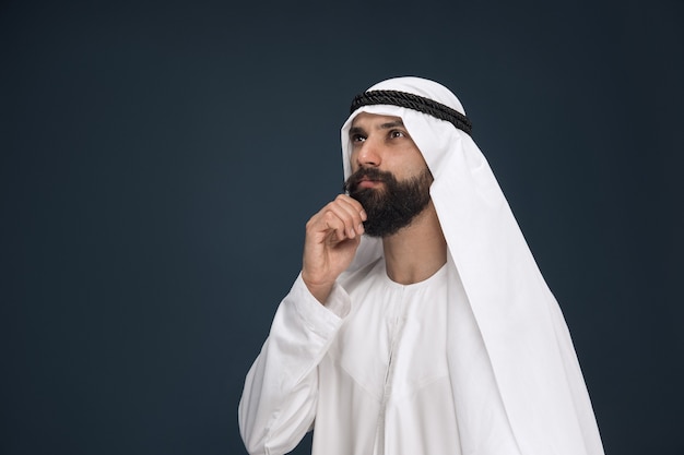 Portrait demi-longueur d'homme d'affaires saoudien arabe sur fond de studio bleu foncé. Jeune modèle masculin debout et a l'air pensif. Concept d'entreprise, finance, expression faciale, émotions humaines.
