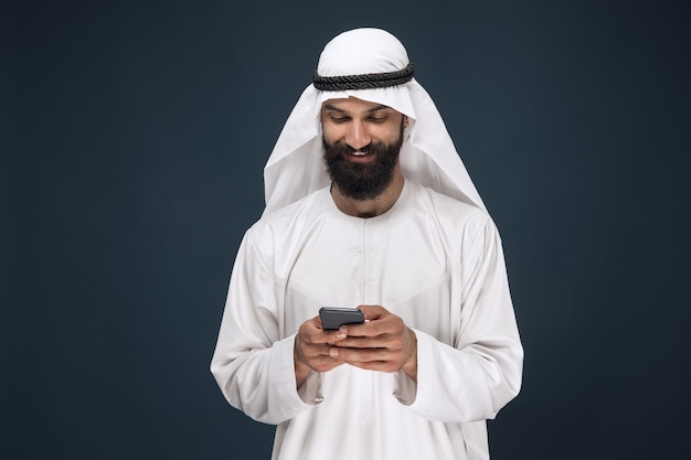 Photo gratuite portrait demi-longueur de l'homme saoudien arabe sur fond de studio bleu foncé. jeune mannequin à l'aide de smartphone, bavarder. concept d'entreprise, finance, expression faciale, émotions humaines, technologies.