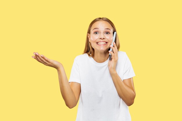 Portrait demi-longueur de la jeune femme caucasienne sur fond de studio jaune. Beau modèle féminin en chemise blanche. Concept d'émotions humaines, expression faciale. Parler au téléphone, souriant.