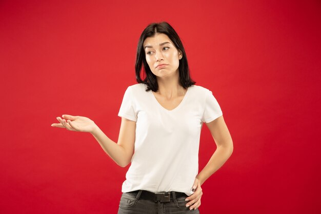 Portrait demi-longueur de la jeune femme caucasienne sur fond de studio rouge. Beau modèle féminin en chemise blanche. Concept d'émotions humaines, expression faciale. Montrer quelque chose d'incertitude, choisir.