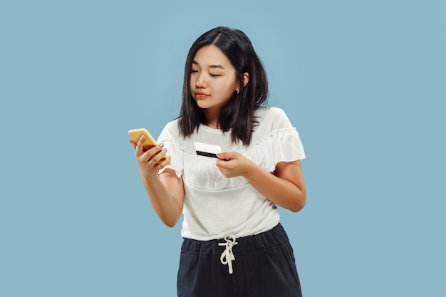 Portrait demi-longueur de la jeune femme coréenne sur l'espace bleu. Modèle féminin utilisant son smartphone pour payer sa facture ou acheter en ligne.