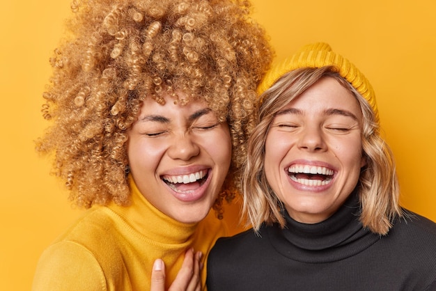 Photo gratuite portrait de deux amies s'amuser rire joyeusement garder les yeux fermés sourire à pleines dents ne pas cacher des sentiments sincères vêtus de cols roulés décontractés isolés sur fond jaune personnes et émotions