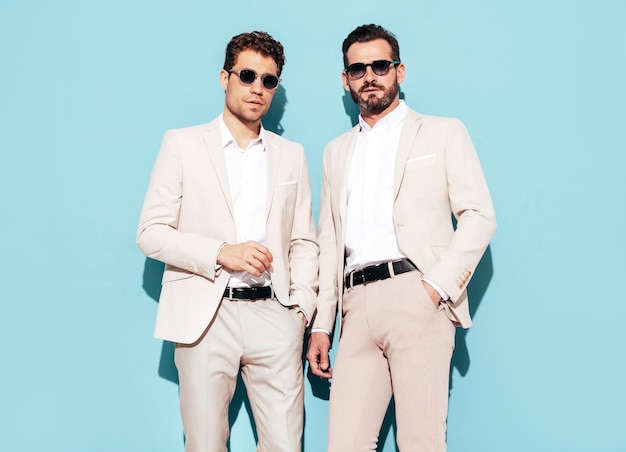 Portrait de deux beaux modèles lambersexuels hipster élégants et confiants Sexy hommes modernes vêtus d'un costume élégant blanc Mode homme posant en studio près du mur bleu en lunettes de soleil