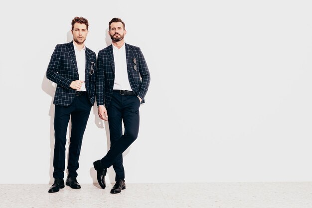 Portrait de deux beaux modèles lambersexuels hipster élégants et confiants Sexy hommes modernes vêtus d'un costume élégant noir Mode homme posant en studio près d'un mur blanc Pleine longueur