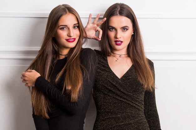 Portrait de deux élégantes jeunes femmes en robes longues glamour posant sur un mur blanc