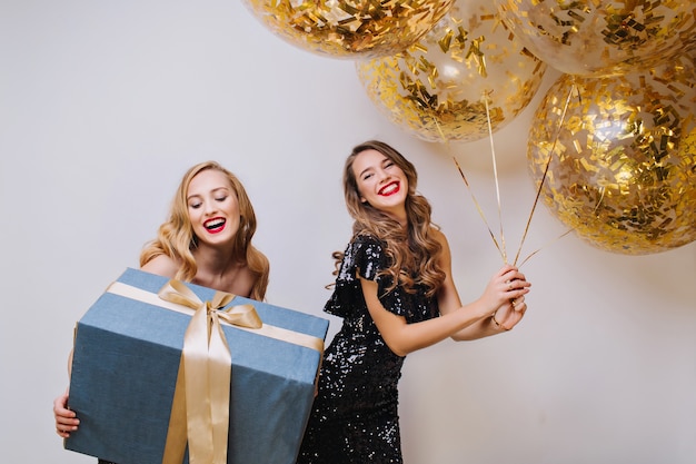 Portrait de deux femmes magnifiques excitées joyeuses avec de longs cheveux bouclés célébrant la fête d'anniversaire sur l'espace blanc. Gros cadeau, ballons avec des guirlandes dorées,