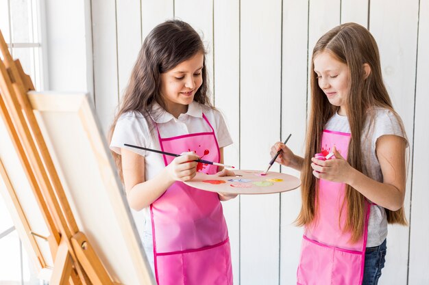 Portrait de deux filles souriantes, debout près du chevalet, mélangeant la peinture avec des pinceaux dans la palette