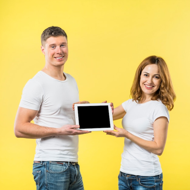 Portrait de deux heureux jeune couple montrant une tablette numérique écran noir sur fond jaune