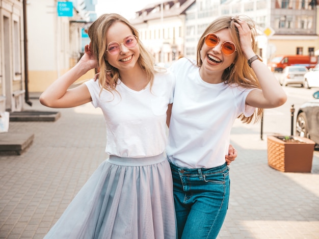 Portrait de deux jeunes belles filles blondes souriantes hipster dans des vêtements de t-shirt blanc à la mode d'été. . Modèles positifs s'amusant avec des lunettes de soleil.