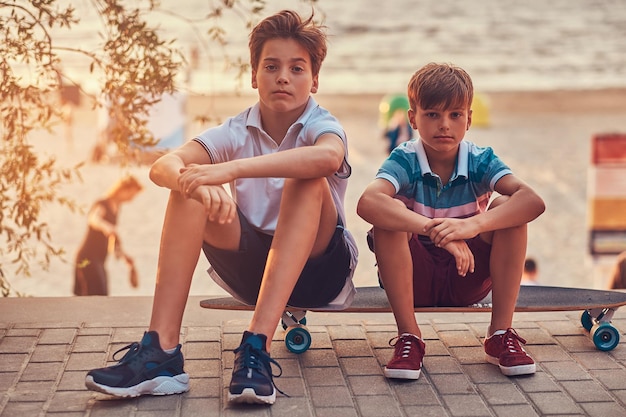 Portrait de deux petits frères assis ensemble sur une planche à roulettes sur fond de côte au coucher du soleil.