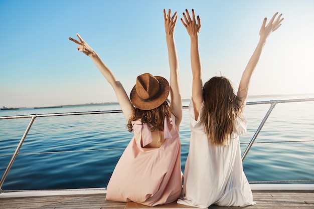 Photo gratuite portrait de dos de deux femelles en robes, assis sur le côté du yacht et en agitant, exprimant le bonheur tout en regardant le bord de mer. qui d'autre peut applaudir mieux qu'un ami proche qui voyage avec vous