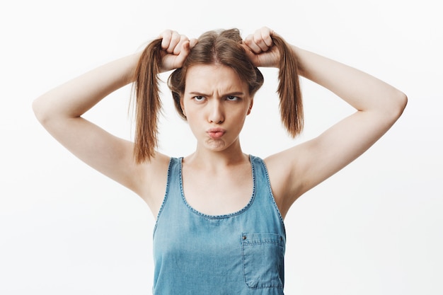 Portrait de drôle belle fille européenne charmante avec des cheveux raides foncés, tondre les yeux, tenant les cheveux avec les deux mains, ayant un look drôle et maladroit. Émotions et expressions positives