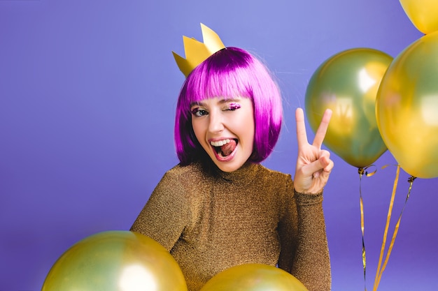 Photo gratuite portrait drôle jeune femme avec coupe de cheveux violet s'amuser. des ballons dorés entourent, montrant la langue, exprimant des émotions positives, une couronne sur la tête, une fête d'anniversaire.