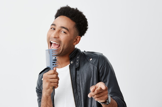 Portrait d'un drôle de jeune homme à la peau noire avec des cheveux bouclés noirs en t-shirt blanc décontracté et une veste en cuir tenant le peigne dans les mains, prétendant qu'il chante avec un microphone tout en se préparant pour la réunion.