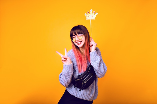 Portrait drôle lumineux de femme joyeuse hipster aux cheveux rose vif, portant un pull confortable, tenant une fausse couronne de fête et souriant, prêt pour la fête, mur jaune.