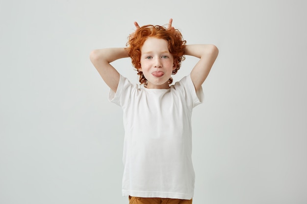 Portrait de drôle petit garçon rousse avec des taches de rousseur s'amuser à l'intérieur, sortir sa langue et faire des cornes avec les doigts.