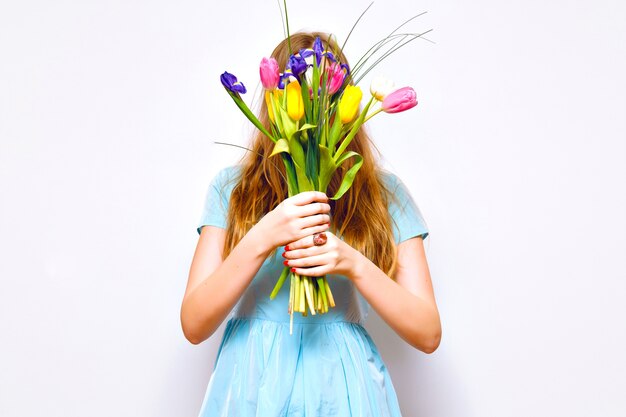 Portrait drôle de studio de femme blonde ferme son visage par beau bouquet de tulipes colorées, couleurs pastel tendres, robe vintage, poils longs, détails de mode. le printemps arrive