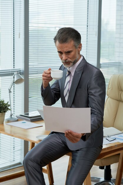 Portrait du puissant PDG perché sur le bureau en train de parcourir le rapport et de siroter un café
