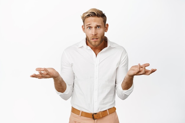 Photo gratuite portrait d'élégant beau mec blond haussant les épaules, l'air confus, ne peut pas comprendre qch debout sans aucune idée avec les mains vides sur le côté fond de studio