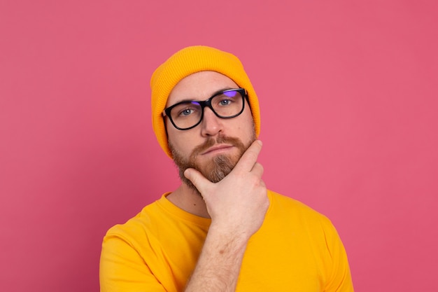 Portrait de l'élégant bel homme barbu européen en chapeau de chemise jaune décontracté et lunettes sur fond rose