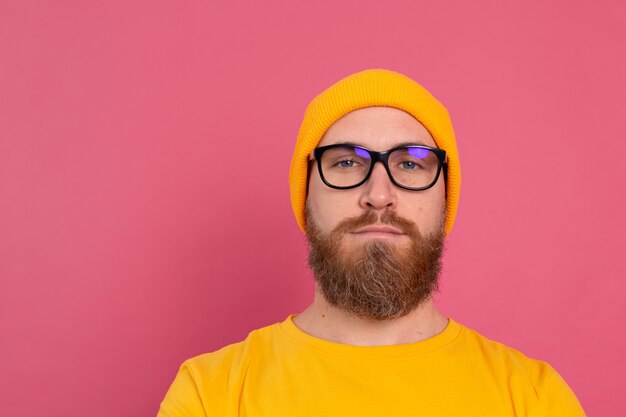 Portrait de l'élégant bel homme barbu européen en chapeau de chemise jaune décontracté et lunettes sur rose