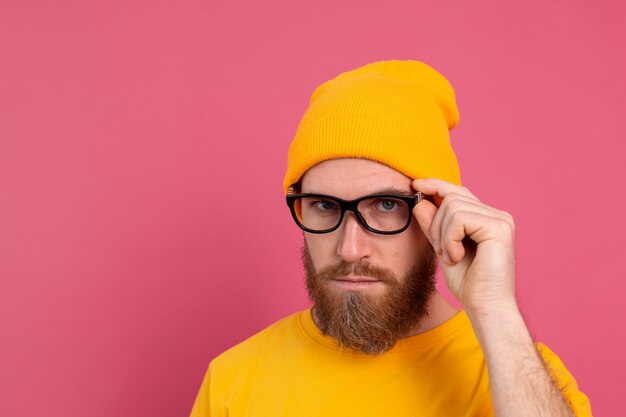 Portrait de l'élégant bel homme barbu européen en chapeau de chemise jaune décontracté et lunettes sur rose