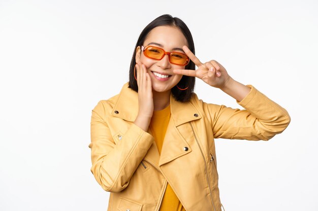 Portrait d'une élégante fille moderne asiatique portant des lunettes de soleil et une veste jaune montrant le geste de vsign de paix debout sur fond blanc visage souriant heureux