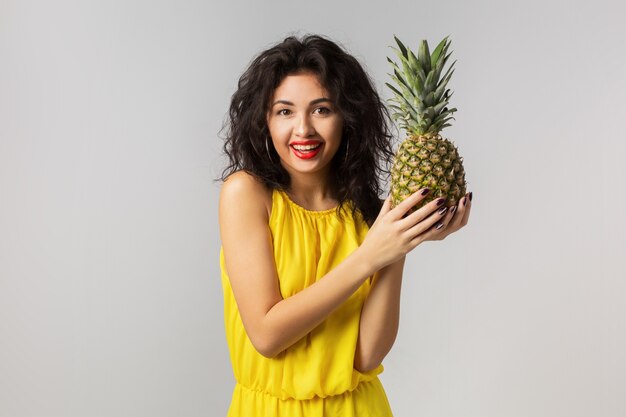 Portrait émotionnel de la jeune femme brune exotique en robe jaune, tenant l'ananas, expression du visage drôle, émotion positive, isolé, fruits tropicaux, alimentation, heureux, souriant, mode de vie sain