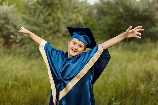 Portrait émotionnel d'une petite étudiante diplômée heureuse dans une robe de graduation bleue avec chapeau, titulaire d'un diplôme, en plein air. écolière gaie chanceuse célébrant le triomphe.