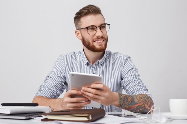 Portrait d'un employé de bureau à lunettes rondes, a des bras tatoués, tient une tablette moderne