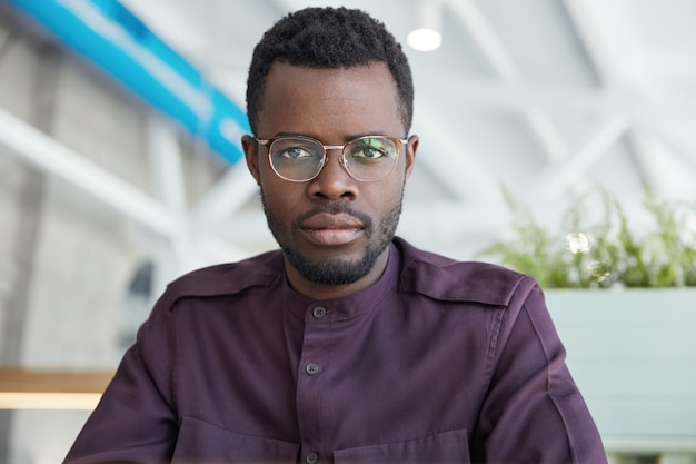 Portrait d'un employé de bureau masculin confiant sérieux en lunettes et chemise formelle, avec une peau foncée, pose dans une armoire spacieuse