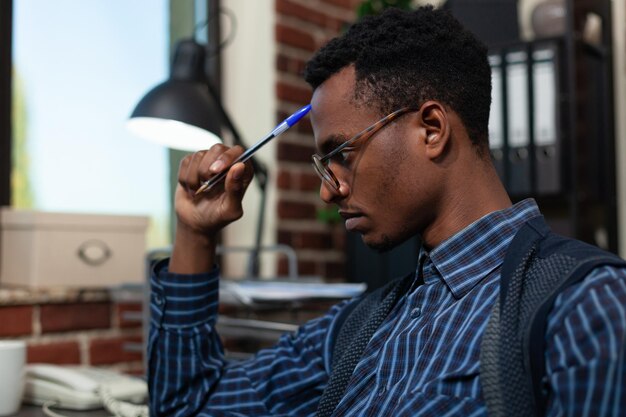 Portrait d'un employé de démarrage touchant le front avec un stylo regardant concentré sur l'écran d'un ordinateur portable avec des analyses d'entreprise. Afro-américain avec des lunettes pensant aux résultats des ventes assis au bureau.