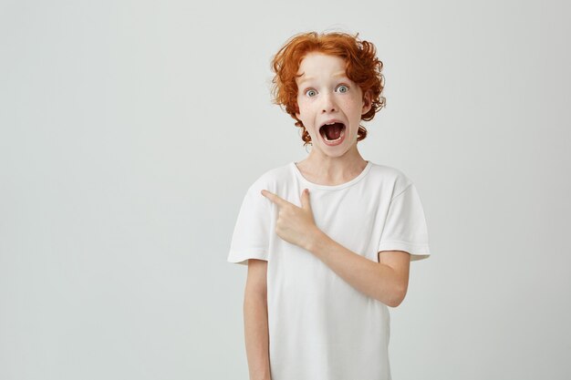 Portrait d'enfant gingembre joyeux avec des taches de rousseur posant avec la bouche ouverte et une expression folle, pointant sur fond