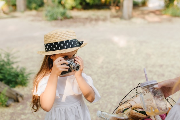 Portrait d'enfant sérieux avec appareil photo porte un chapeau de canotier à la mode décoré d'un ruban noir. Petite fille aux cheveux bruns prenant la photo du panier de pique-nique tenant sa mère.
