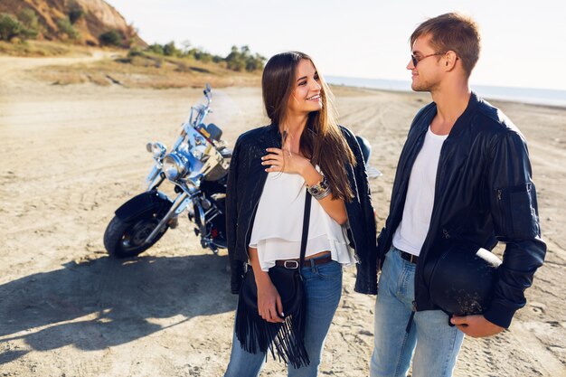 Portrait ensoleillé de mode de vie de cavaliers de jeune couple posant ensemble sur la plage en moto - concept de voyage. Deux personnes et vélo. Image de mode d'une femme et d'un homme sexy étonnants parlent et rient.