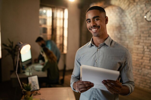 Portrait d'un entrepreneur noir heureux travaillant tard sur des rapports d'activité et regardant la caméra au bureau