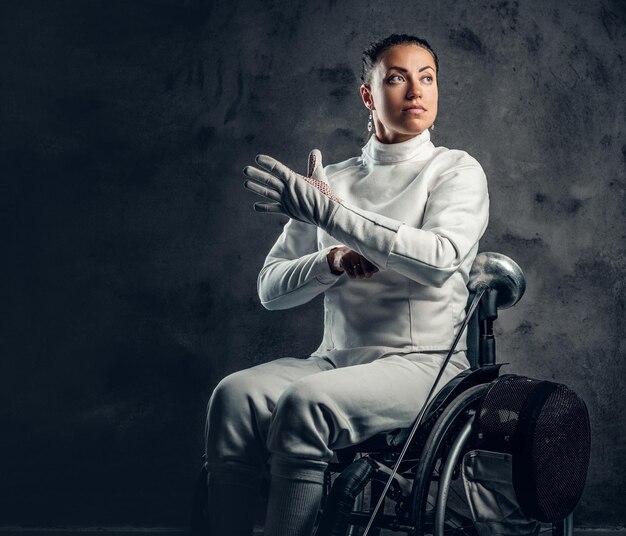 Portrait d'une escrimeuse en fauteuil roulant avec masque de sécurité et rapière sur fond gris.