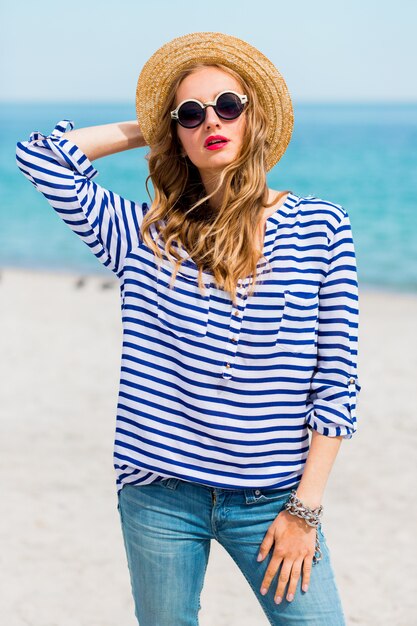 Portrait d'été de jeune femme fraîche dans des lunettes de soleil cool et chapeau de paille posant sur la plage tropicale ensoleillée