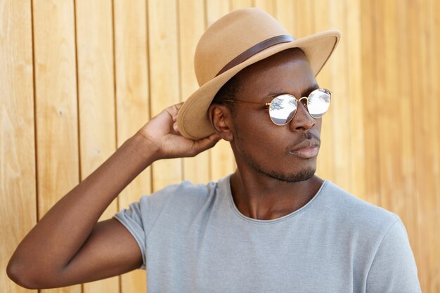 Portrait d'été en plein air de jeune étudiant noir sérieux et confiant se détendre à l'extérieur, ajustant son chapeau élégant, ayant un regard réfléchi