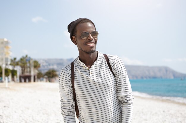 Portrait d'été en plein air de sourire heureux homme à la peau sombre dans des chapeaux à la mode et des nuances de passer une journée ensoleillée sur la plage de la ville, marcher, s'amuser et profiter de la mer