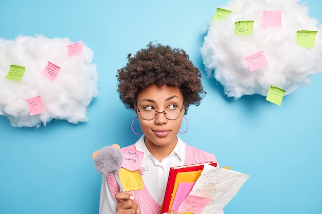 Photo gratuite portrait d'étudiante afro-américaine songeuse détient des dossiers de papiers et un stylo prend des notes pendant la conférence à l'université apprend le matériel pour les poses d'examen contre les nuages avec des autocollants de rappel autour